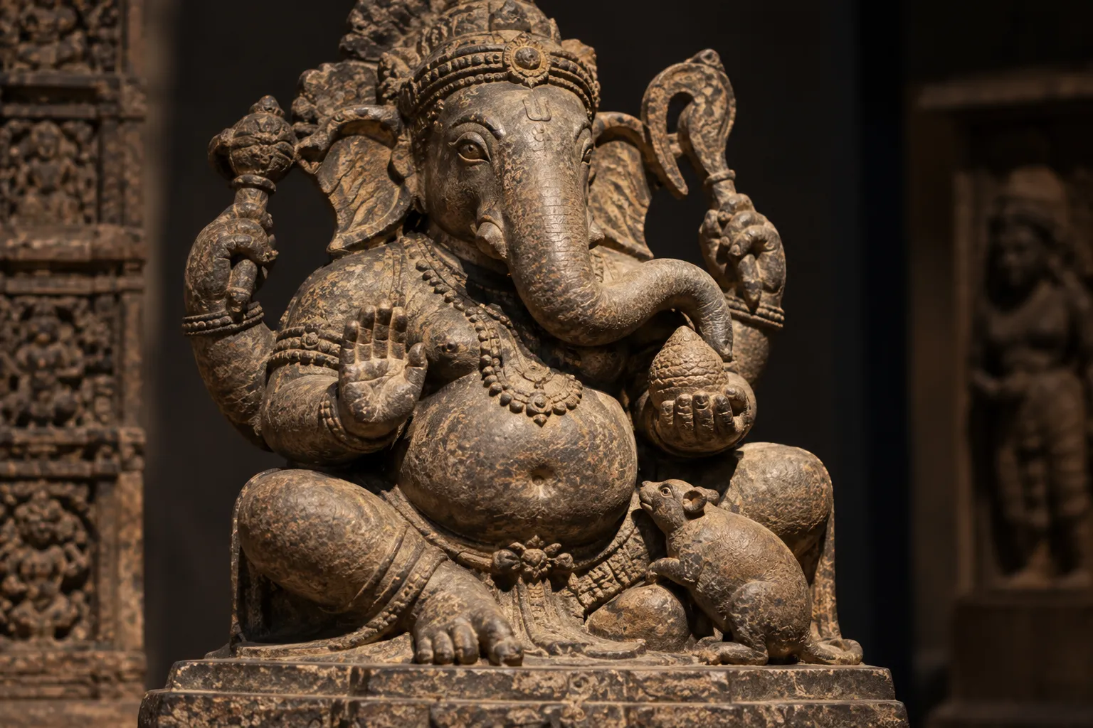 A weathered stone sculpture of Ganesha seated with a small mouse beside his feet, shown in close detail against an ancient temple interior.