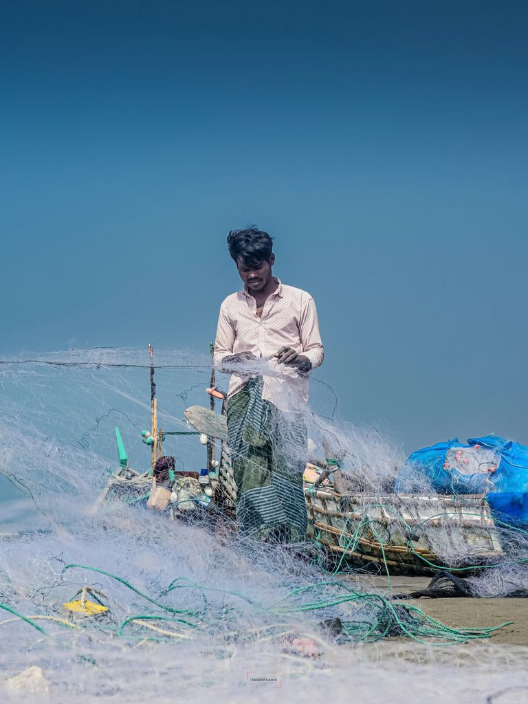 A fisherman repairs his net aboard a wooden boat, under clear blue skies, showcasing rural life.