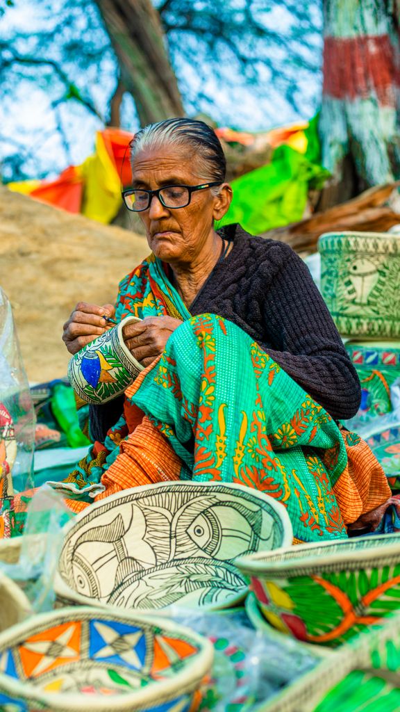 An elderly woman creating vibrant traditional Indian handicrafts in a market setting.